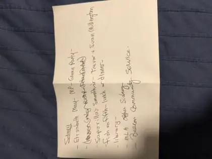 A folded cream note lies sideways on navy quilted fabric, a handwritten list under 'Sidney' reading 'Elizabeth May – MP – Green Party', a crossed-out line, a smoothie shop, 'Fish on Fifth – lunch or dinner', library, 'MLA Office Sidney' and 'Beacon Community Service.'