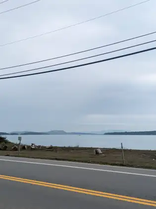 A shoreline road looking across calm water to low islands and distant hills, power lines overhead.
