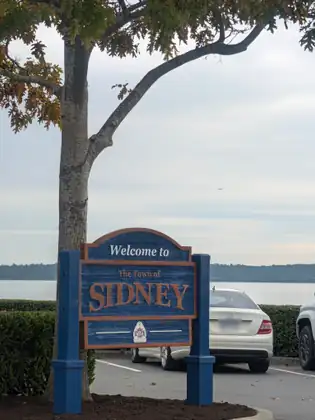 A blue and orange carved sign reading Welcome to the Town of Sidney, parked cars and the water behind it.