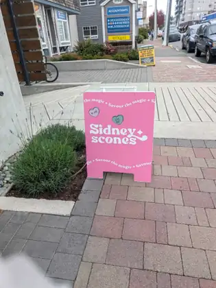A pink Sidney Scones sandwich board on a brick sidewalk, with shops and parked cars along the street behind.