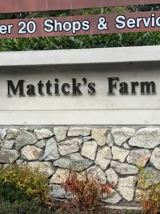 The words Mattick’s Farm in raised dark letters on a pale concrete band above a stone wall, with part of a brown sign about its shops and services above.