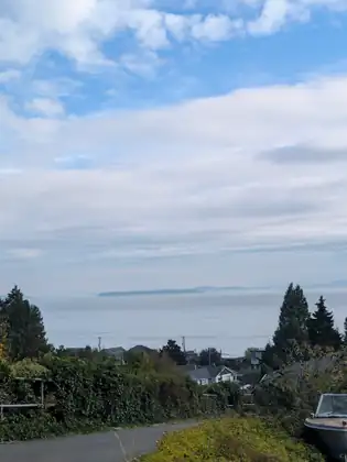 A paved lane running downhill past hedges toward rooftops, the bow of a parked boat at the edge, with the sea, a long low island and distant hills beyond under blue sky and white cloud.
