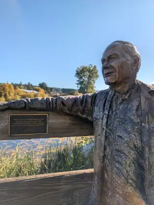 A weathered bronze statue of an older man in an open-collared shirt and jacket, one arm resting along a wooden railing, beside a plaque from Mayor Frank Leonard and Council honouring the early settlers of the Blenkinsop Valley, reeds and water below.