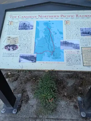 An interpretive sign titled The Canadian Northern Pacific Railway, with old photographs of a railcar, a locomotive and trestle bridges and a map of historical railways through Saanich, weeds growing up through the concrete beneath it.