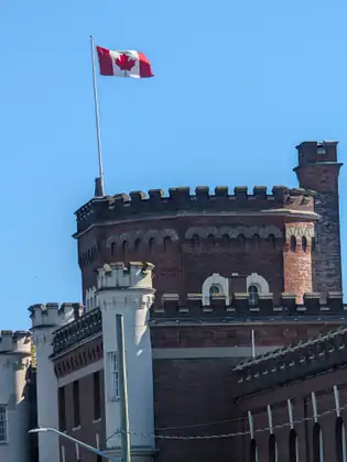 A red-brick building with castle-like crenellated towers and pale turrets, the Canadian flag flying above it against a clear blue sky.