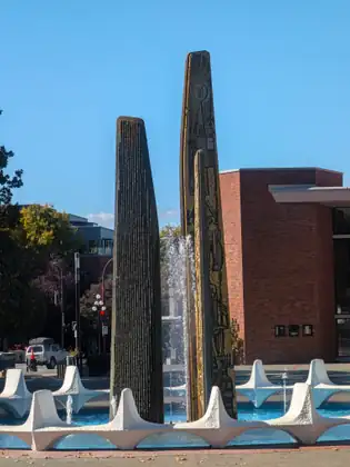 A fountain with tall, narrow columns, one ribbed and one decorated with figures and symbols, spraying water into a blue basin ringed with white, wave-like concrete forms, a red-brick building to the right.