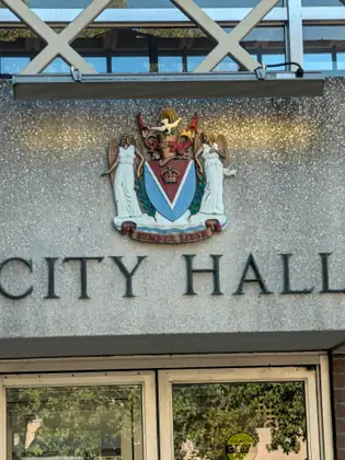The words City Hall in dark raised letters on a speckled concrete wall above glass doors, beneath a painted coat of arms with two winged figures, a crown on a red and blue shield and the motto Semper Liber.