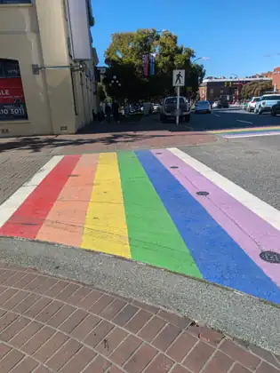 A rainbow-striped crosswalk at a downtown intersection, brick sidewalks on either side, with people walking past a cream-coloured corner building, a pickup and cars on the street and red-brick buildings beyond under a blue sky.