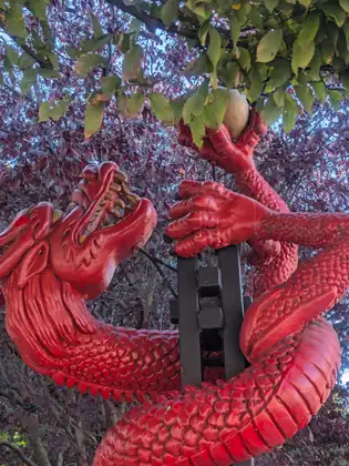 A red dragon sculpture coiled around a dark post, its jaws open and one claw raised to grasp a pale ball, under a canopy of green and purple leaves.