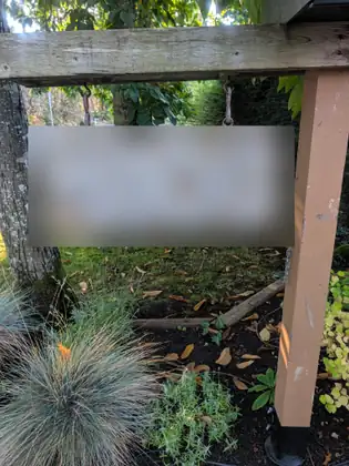 A weathered wooden house-number sign, its numbers blurred, hangs on chains from a wooden crossbeam above a garden bed of blue-gray ornamental grass, fallen leaves and small green plants.