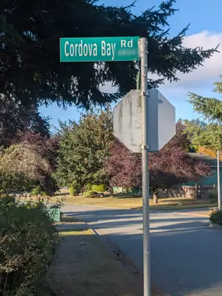 A green street sign reading Cordova Bay Rd, its block number blurred, tops a metal post that also holds the back of a stop sign, at a quiet corner with a curving road, lawns, purple-leaved trees and a green house.