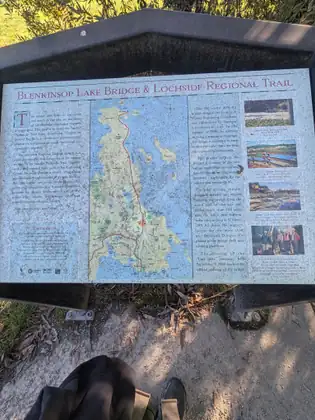 An interpretive sign titled Blenkinsop Lake Bridge & Lochside Regional Trail, with a map tracing the trail’s route and photographs of the bridge and its construction, a black pack with tan straps and the toe of a shoe at the bottom of the frame.