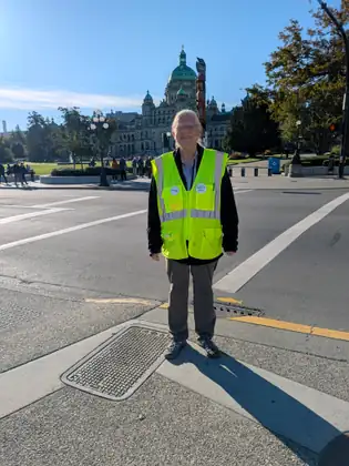 A smiling white-haired woman in a high-visibility vest with an NMDP button and a HERO button pinned to it stands on a street corner, with the green-domed BC Legislature and a tall carved totem pole behind her.