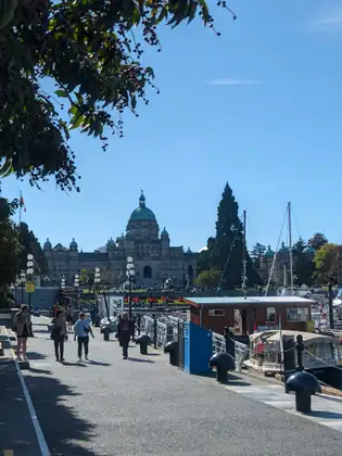 People stroll toward the camera along the Inner Harbour walkway, which leads to the green-domed BC Legislature, past black bollards, a small wooden dock hut and covered harbour boats moored on the right.
