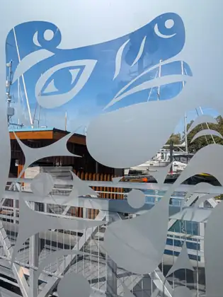 A frosted-glass panel cut with bold, stylised curving shapes and large eye forms frames a view of a dock gangway, a wooden harbour hut and sailboat masts beyond.