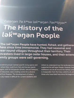 An interpretive sign headed 'The History of the lək̓ʷəŋən People', with a line in the lək̓ʷəŋən people's own language above, says they have hunted, fished and gathered here since time immemorial, and that their ancestors lived in large cedar houses.