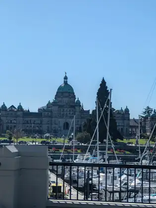 Beyond a black railing and a pale concrete post, sailboat masts and moored boats fill the marina below the green-domed BC Legislature, its long stone facade and smaller domed towers stretching across under a clear blue sky.