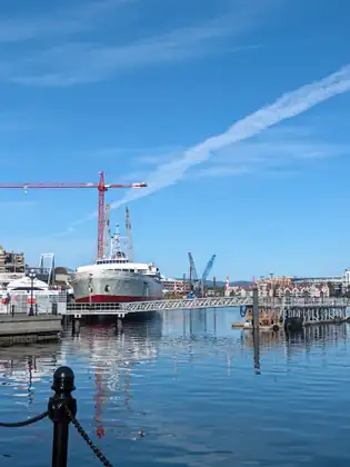 A white ship with a red band along the bottom of its hull sits moored behind a long metal gangway on the still Inner Harbour, with an FRS Clipper terminal sign on the left and red and blue construction cranes behind.