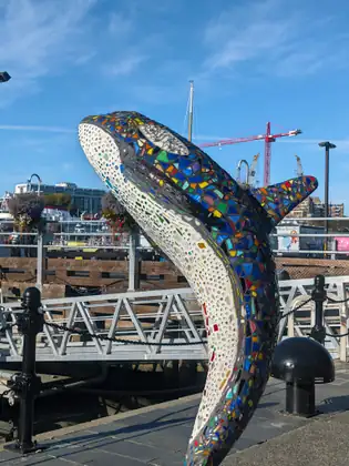A sculpture of a leaping whale, covered in colourful broken-tile mosaic, rises from the harbour walkway beside a dock railing, with a red construction crane in the distance.