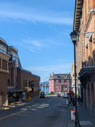 A quiet street runs down between brick buildings to the blue water of the harbour, ending at a pink two-storey building with a slate mansard roof and distant hills beyond.