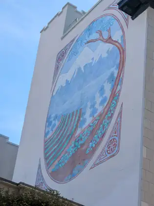 A large mural on a white building wall shows, inside an oval frame, a red-brown tree trunk arching over striped fields, blue trees and snow-capped mountains.