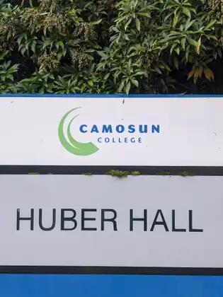 A white sign with the green crescent Camosun College logo above the words Huber Hall, with a leafy shrub growing over its top edge.