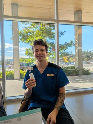 A smiling young man in blue nursing scrubs holds up a pair of barcoded cheek swabs in their paper sleeve, in front of tall windows looking out on a tree and a parking lot.