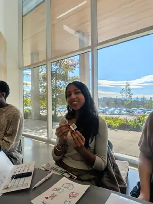 A smiling young woman holds up a cheek swab with its barcode label, seated at a table of swab instructions in front of tall windows overlooking trees and a parking lot.