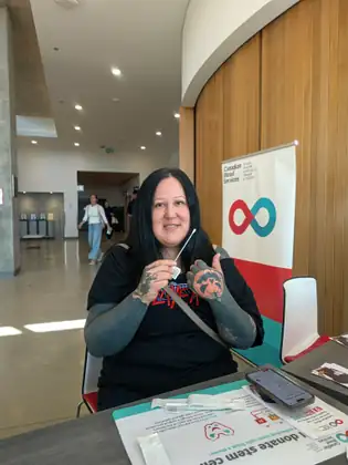 A smiling woman with tattooed arms and hands holds up a cheek swab at a table beside a Canadian Blood Services banner reading Blood, Plasma, Stem Cells, Organs and Tissues.