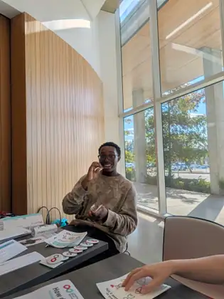 Laughing, a young man in glasses rubs a cheek swab inside his mouth at a table spread with #GetSwabbed leaflets and red-and-teal infinity-logo buttons.