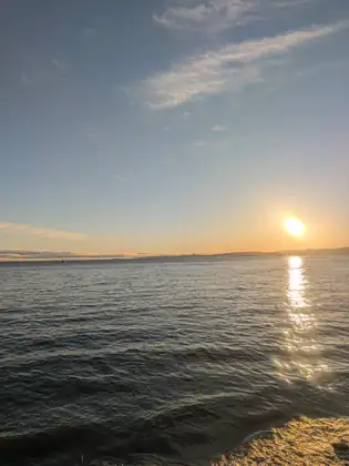 The low sun hangs just above a long line of distant hills over a calm sea off Victoria, laying a glittering path of light across the water toward the sunlit rocky shore in the foreground.