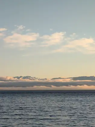 A band of low cloud lies over the sea at dusk, with jagged mountain peaks rising above it under a pale peach-and-blue sky.