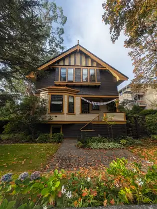 A dark-shingled house with cream siding and mustard-yellow trim, a string of small pale flags across its porch, stands behind a leaf-strewn paved path, a lawn, blue and purple flower heads and white daisy-like flowers under tall trees.