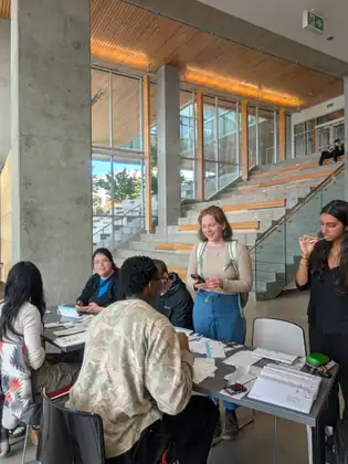 Students gather around a table spread with swab kits and envelopes in a bright concrete-and-timber atrium, while a young woman on the right runs a cheek swab inside her mouth.