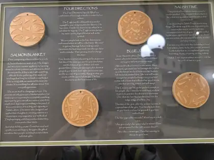 A framed display of four carved wooden discs with Salish designs, titled Salmon Blanket, Four Directions, Blue Jay and Salish Time, each beside a block of text explaining its meaning.