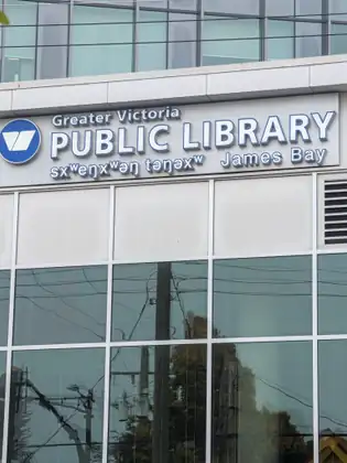 The Greater Victoria Public Library James Bay sign, with a second line in another language, sits above tall glass windows reflecting power lines and trees.