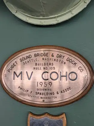 A copper builder's plaque on a green bulkhead reads M.V. Coho, 1959, Hull No. 105, built by Puget Sound Bridge & Dry Dock Co. of Seattle and designed by Philip F. Spaulding & Assoc.