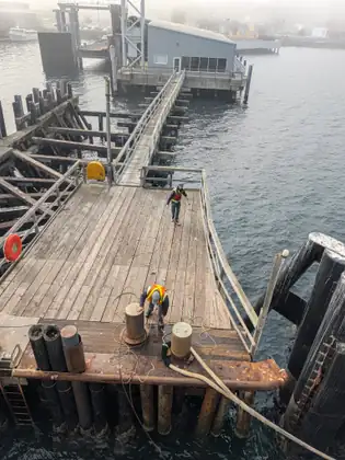 Seen from the ferry above, two workers in high-visibility vests handle mooring lines on a weathered wooden dock, with a gangway leading to a grey terminal building over misty water.