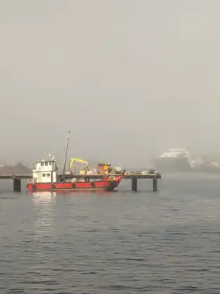 A red work boat with a white cabin is tied up at a low pier where a yellow crane sits among equipment, while a large ship looms in the fog behind.