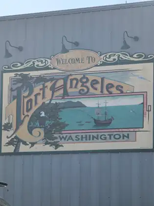 A painted Welcome to Port Angeles, Washington sign on a corrugated metal wall shows an eagle perched in a tree beside a tall sailing ship and small boats in a blue bay below mountains.