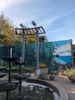 A tiered fountain splashes in a brick plaza in front of a long mural of tall forest, a rock standing offshore and ocean surf, under a blue sky with autumn trees above.