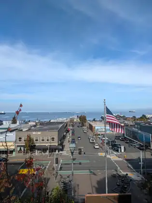 From a raised viewpoint with an American flag, a downtown Port Angeles street runs straight down to the harbour, where large ships sit on the blue water under a wide sky.