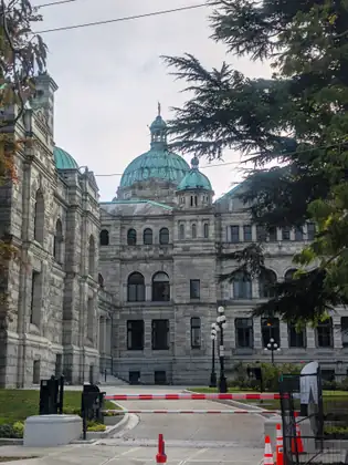 The grey stone BC Legislature, with green copper domes and a small gilded figure on top, seen past two red-and-white gate barrier arms, traffic cones and a large evergreen.