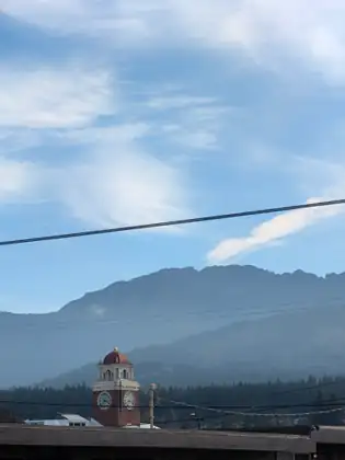 A brick clock tower with a red dome rises above rooftops and dark forest, with a long, hazy mountain ridge behind under wispy clouds.