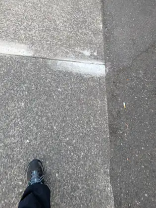 Looking down at a dark hiking shoe mid-step on a pale concrete sidewalk at the edge of the asphalt road.