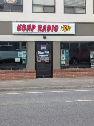The storefront of KONP Radio 1450, with a red-lettered sign above a glass door with station stickers, a low brick base, and dark windows with a few local event flyers taped up.