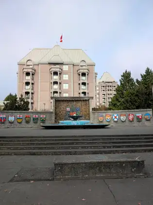 A fountain stands against a low stone wall lined with painted provincial crests, with wide steps and a stone block marked 1967 – 2067 in front and a tall pink building flying a Canadian flag behind.