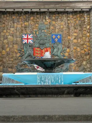 Water spills from a dark bowl in front of a bronze lion and unicorn flanking a coat of arms, with a Union Jack and a blue flag with gold fleurs-de-lis above and water trickling down the stone wall behind.