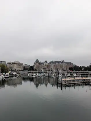 Victoria's Inner Harbour under heavy grey cloud, with sailboats at the docks and a large château-style building with steep roofs reflected in the still water.