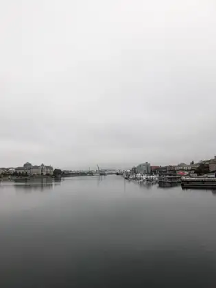 Calm grey water stretches up Victoria's harbour toward a white bridge and construction cranes in the far distance, with a large multi-storey building on the left shore and a marina of moored boats below city buildings on the right.